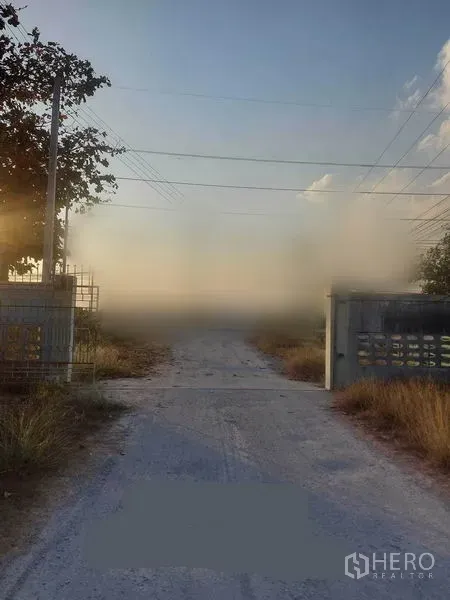 Factory for rent in Mueang Nakhon Pathom, Nakhon Pathom - Gated entrance with concrete access road and power lines leading to the industrial site at dusk.