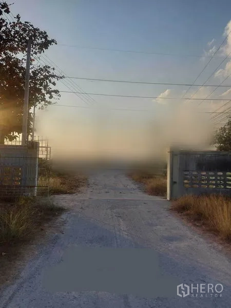 Factory for rent in Mueang Nakhon Pathom, Nakhon Pathom - Gated entrance with concrete access road and power lines leading to the industrial site at dusk.