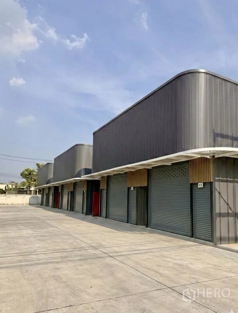 Warehouse for rent in Suan Luang, Bangkok - Row of warehouse units with canopies and rolling doors along a wide concrete driveway.