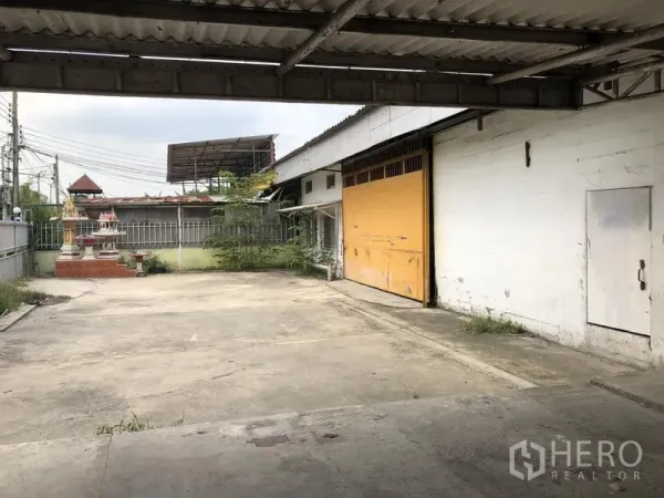 Warehouse for rent in Sai Mai, Bangkok - Gated concrete yard with covered loading bay and a wide yellow roller door at a Sai Mai warehouse.