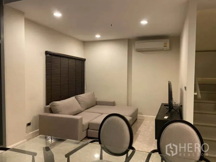 Condominium for rent in Khlong Toei, Bangkok - Open-plan living and dining area with L-shaped sofa, TV console and adjacent staircase.