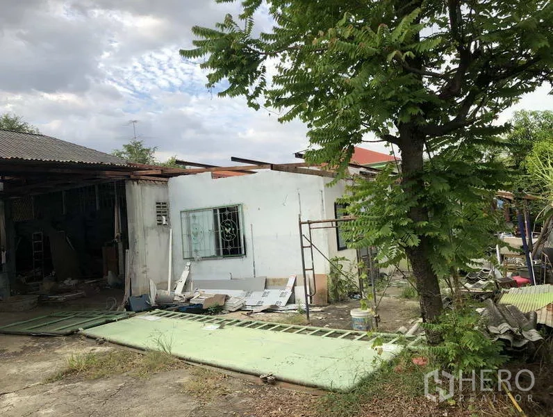 货栈 出租在Sai Mai, Bangkok - 仓库侧面，白色附楼、绿树与散落的建材。