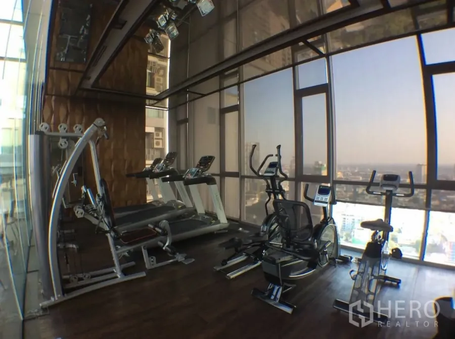 Condominium for rent in Khlong Toei, Bangkok - High-rise condo fitness center with treadmills and bikes facing floor-to-ceiling windows and a Bangkok skyline view.