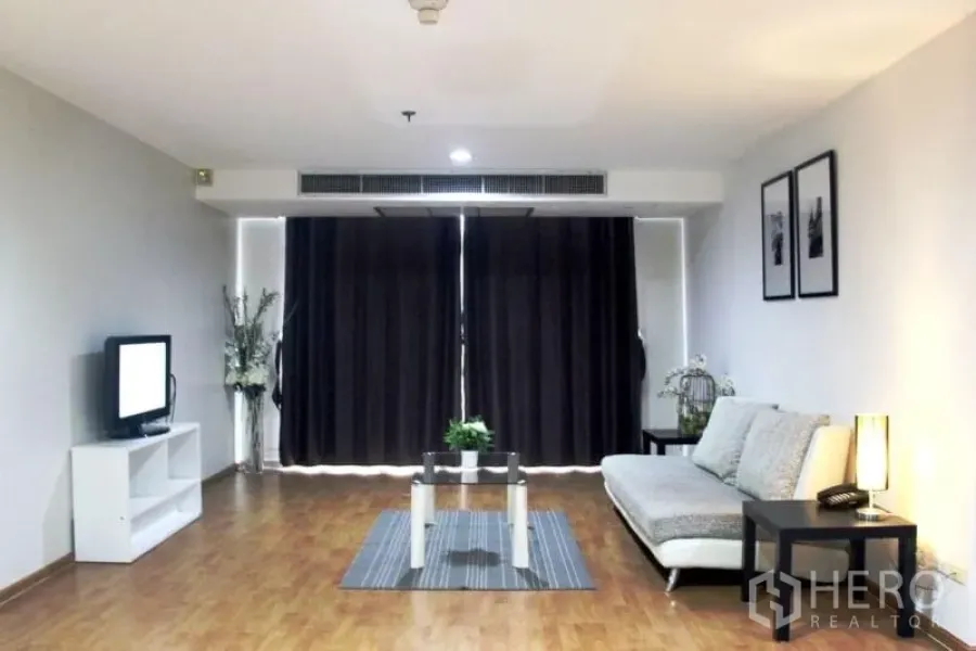 Condominium for rent in Khlong Toei, Bangkok - Living room facing a large window wall with dark drapes, sofa, TV stand and coffee table.