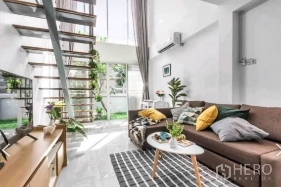Townhouse for rent in Khlong Toei, Bangkok - Airy living room with high ceilings, sectional sofa, and open-tread stairs beside a glass wall to greenery.