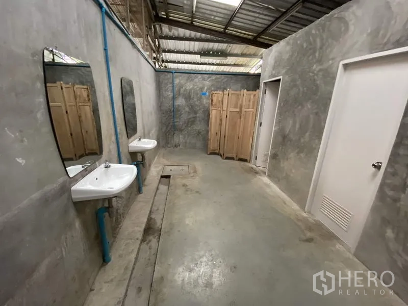 Warehouse for rent or sale in Khlong Luang, Pathum Thani - Industrial restroom with concrete walls, twin sinks, mirrors, and stall doors under a metal roof.