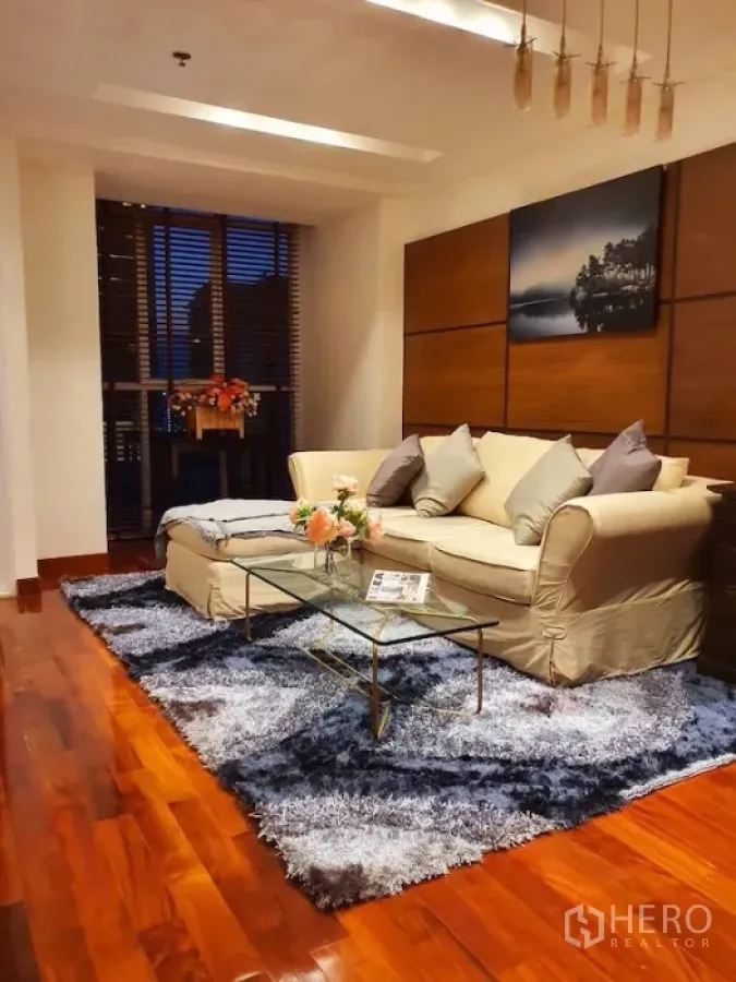 Condominium for rent or sale in Pathum Wan, Bangkok - Warm living room with cream sofa, glass coffee table, plush rug and wooden floors beside a window with blinds.