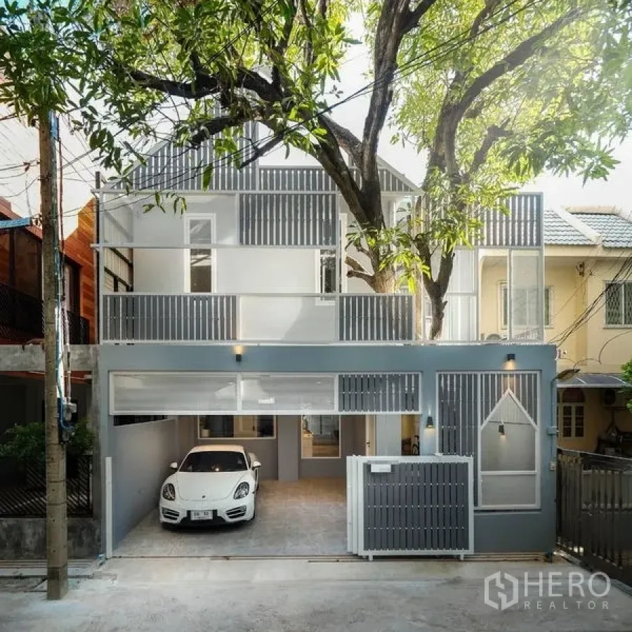 Townhouse for rent in Watthana, Bangkok - Modern townhouse exterior with gray facade, balconies, sliding gate and carport shaded by a tree.