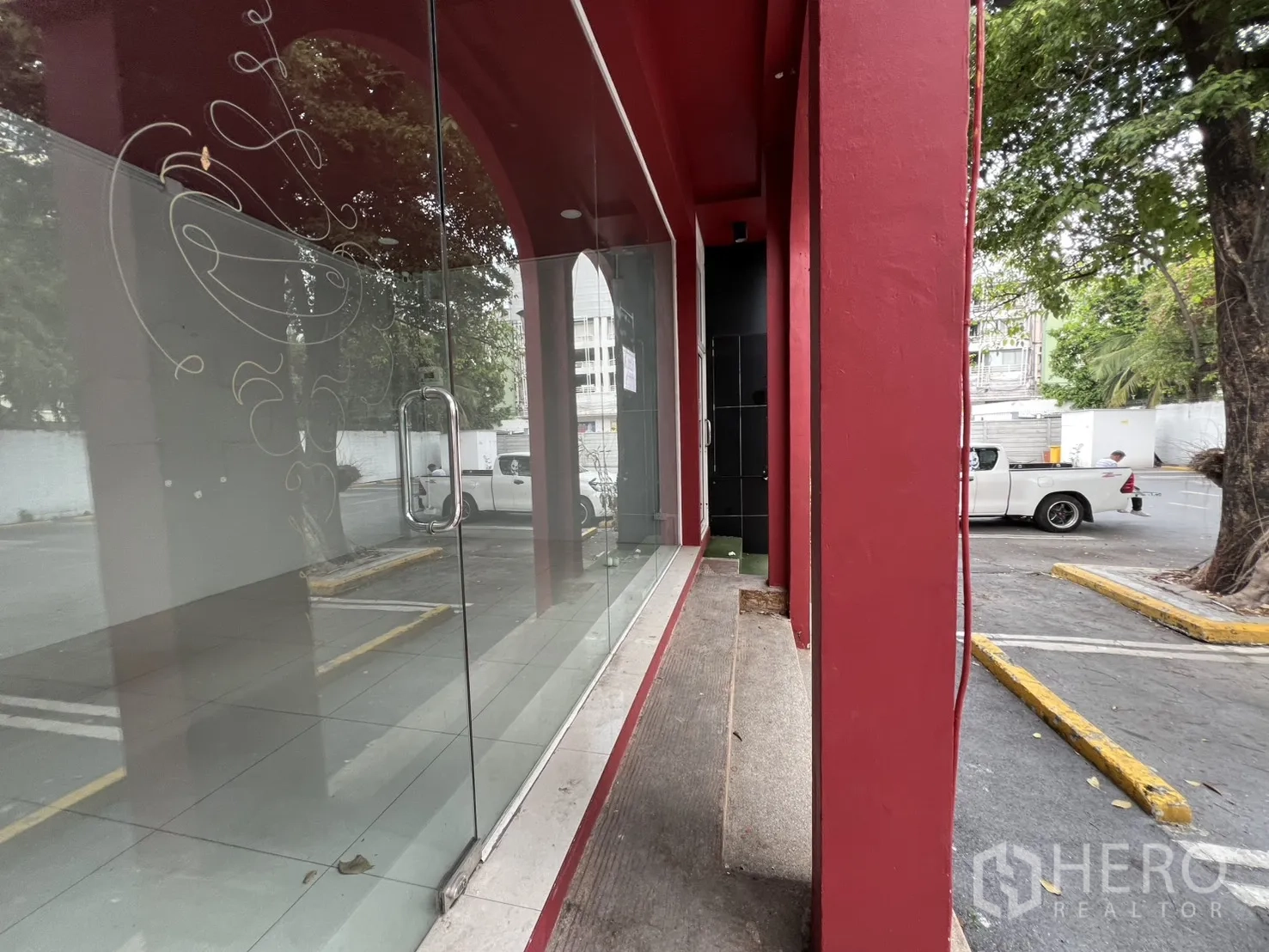 Retail Space for rent in Watthana, Bangkok - Close-up of glass storefront with red columns and pull handle overlooking the parking area and trees.