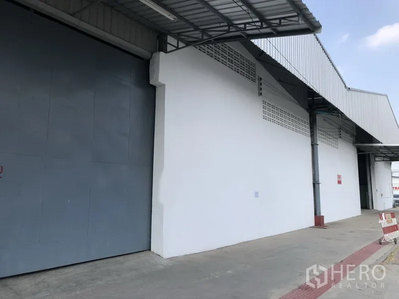 Warehouse for rent in Nonthaburi - Exterior of the warehouse with a large gray sliding door and covered loading bay along a wide driveway.