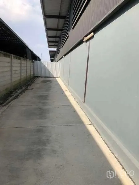 Warehouse for rent in Bang Phli, Samut Prakan - Side driveway with concrete paving and boundary wall running along the warehouse.
