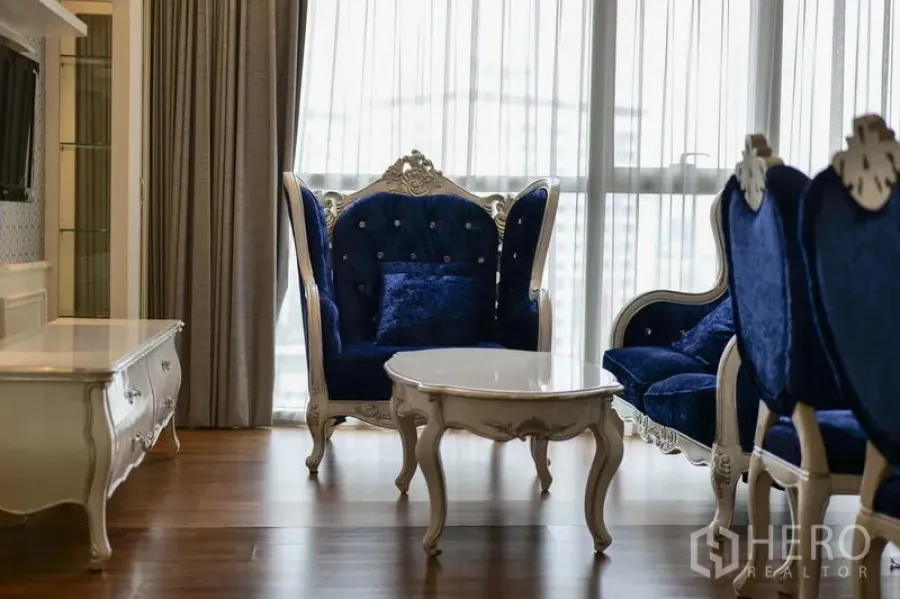 Condominium for rent in Khlong Toei, Bangkok - Elegant lounge with blue velvet armchairs and sofa around a white coffee table by full-height windows.