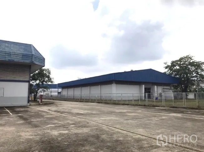 Factory for rent in Bang Bua Thong, Nonthaburi - Open concrete yard between blue-roof warehouses and perimeter fence.