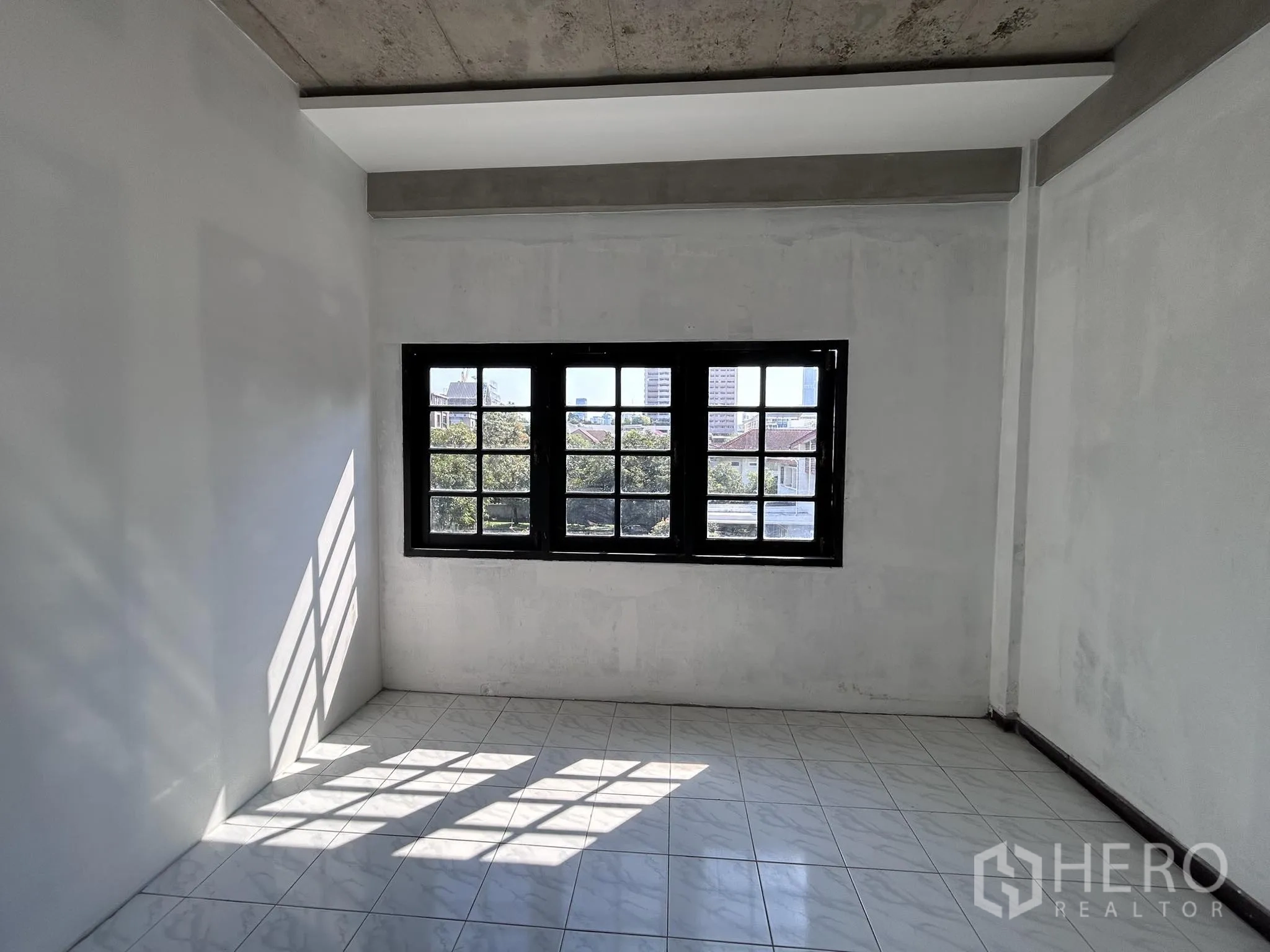 Home Office for rent or sale in Watthana, Bangkok - Sunlit tiled room with black-framed window and exposed concrete beams.