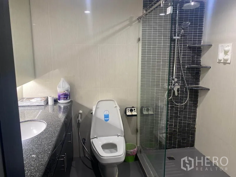 Condominium for rent in Khlong Toei, Bangkok - Bathroom with glass-enclosed shower, rainfall head, mosaic tile wall, and toilet.