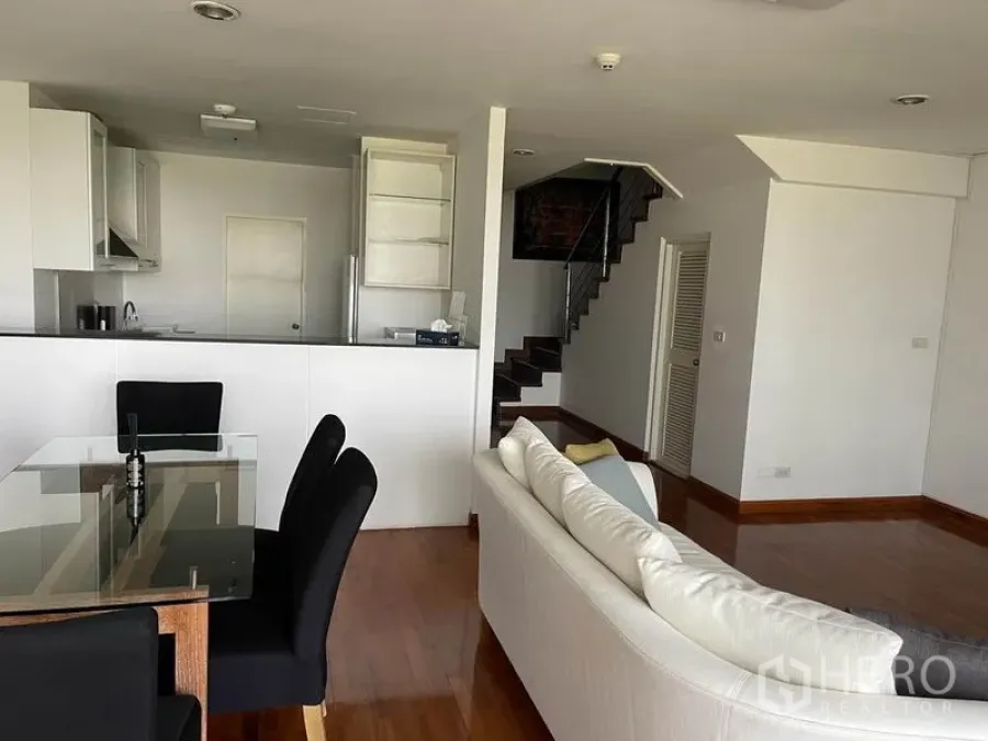 Condominium for rent in Pathum Wan, Bangkok - Open-plan Bangkok condo living area with glass dining table, white sofa, and a compact kitchen beside a staircase.