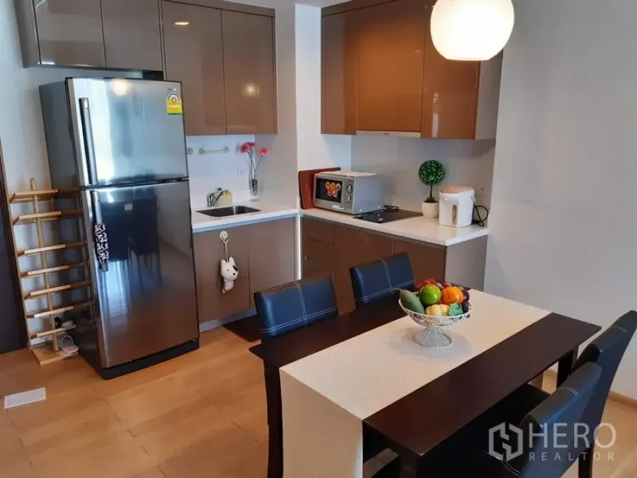 Condominium for rent in Khlong Toei, Bangkok - Modern L-shaped kitchen with glossy cabinets, stainless refrigerator, microwave, and a four-seat dining table in a 1-bedroom Sukhumvit condo.