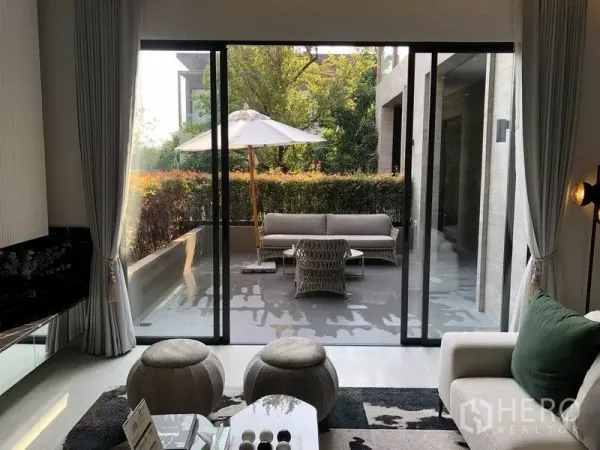 Detached House for sale in Bang Kapi, Bangkok - Living room with sliding glass opening to a shaded terrace with outdoor sofa, umbrella and hedge garden.