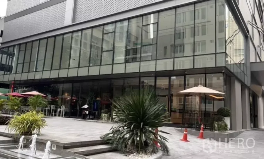 Retail Space for rent - Modern retail building with glass facade, patio umbrellas, plants and a small fountain.