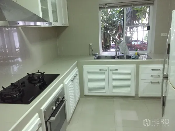 Detached House for rent in Watthana, Bangkok - U-shaped built-in kitchen with gas hob, oven, stainless hood, double sink and window view.