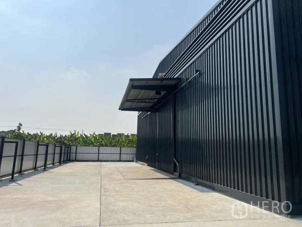 Warehouse for rent in Lat Krabang, Bangkok - Fenced concrete yard beside a black steel warehouse, ample truck turning space and canopy.