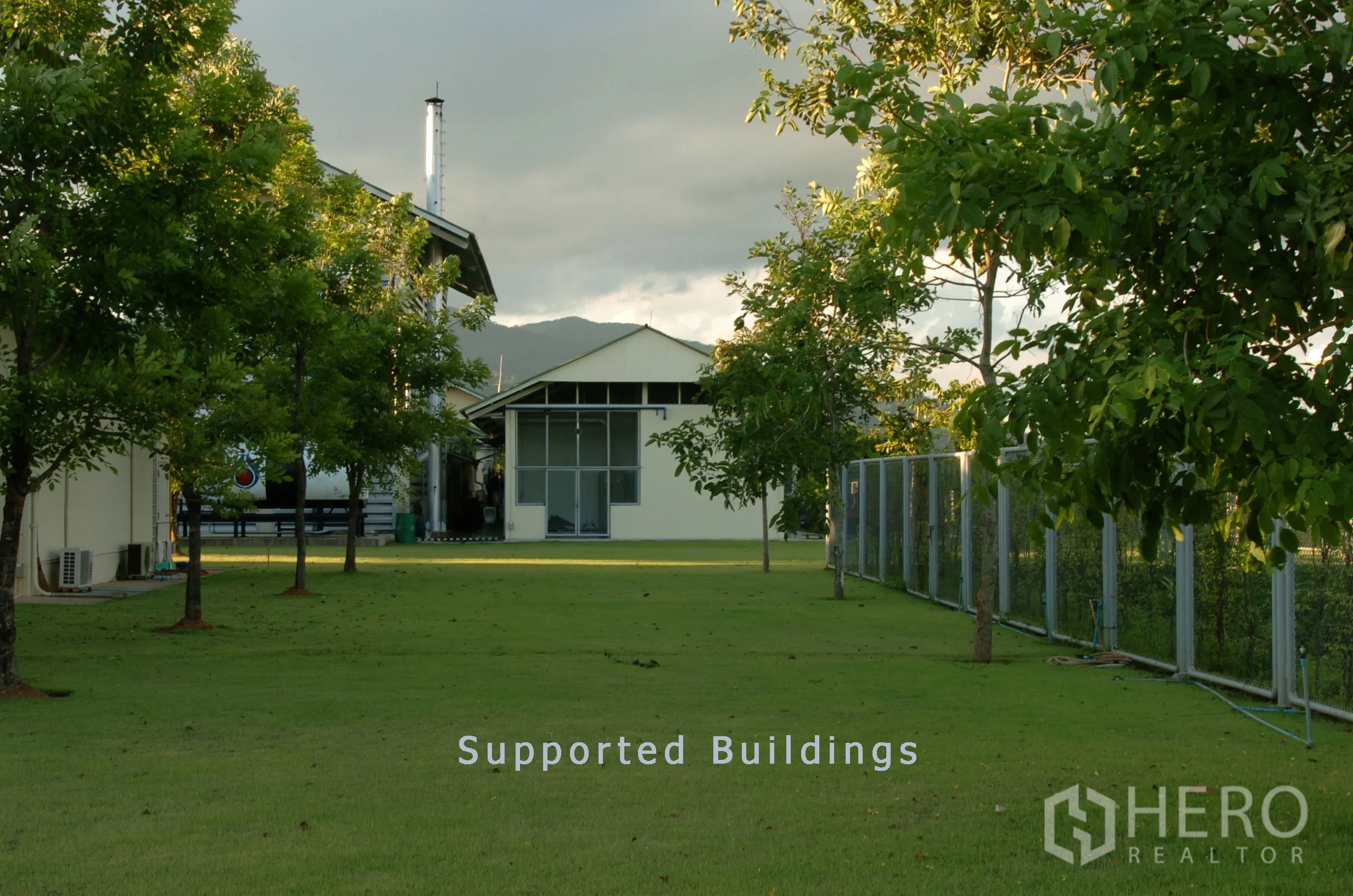 Factory for sale in Khao Chamao, Rayong - Support buildings set behind a green lawn and perimeter fence with hills in the distance.