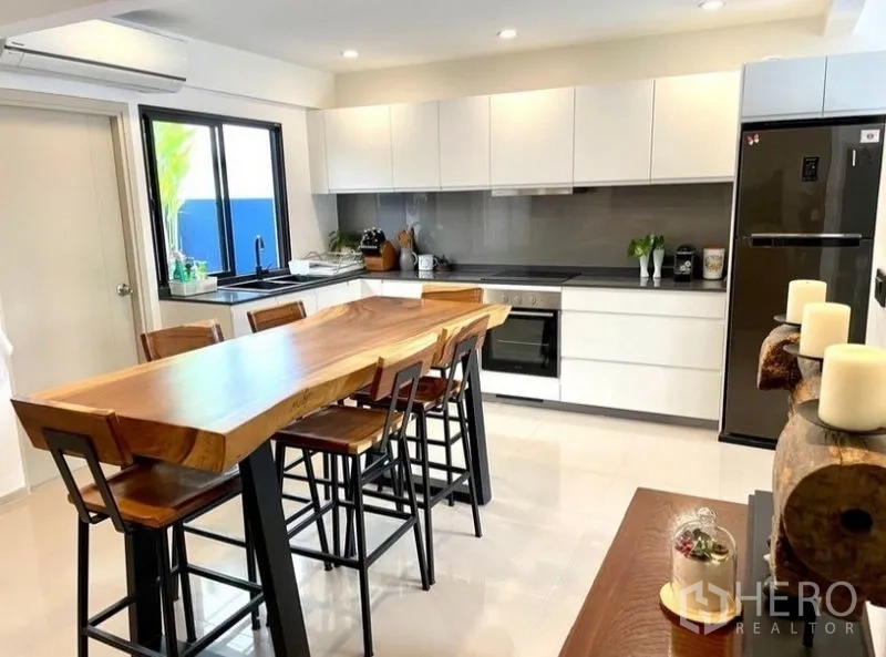 Townhouse for rent in Suan Luang, Bangkok - Contemporary white kitchen with a long solid-wood island dining table and built-in appliances.