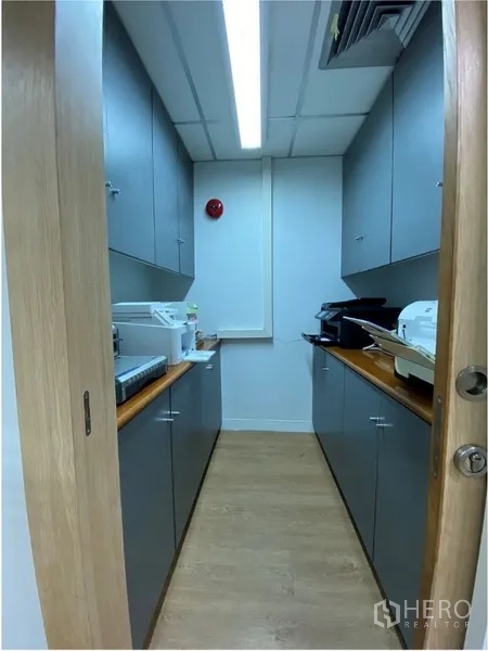Office Space for rent in Khlong Toei, Bangkok - Compact office utility room with grey cabinets, wood floors, built-in countertops and printers under bright fluorescent lighting.