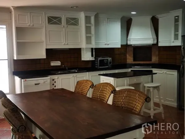 Detached House for rent in Mueang Samut Prakan, Samut Prakan - Country-style kitchen with white cabinetry, center island and a long wooden dining table with rattan chairs.