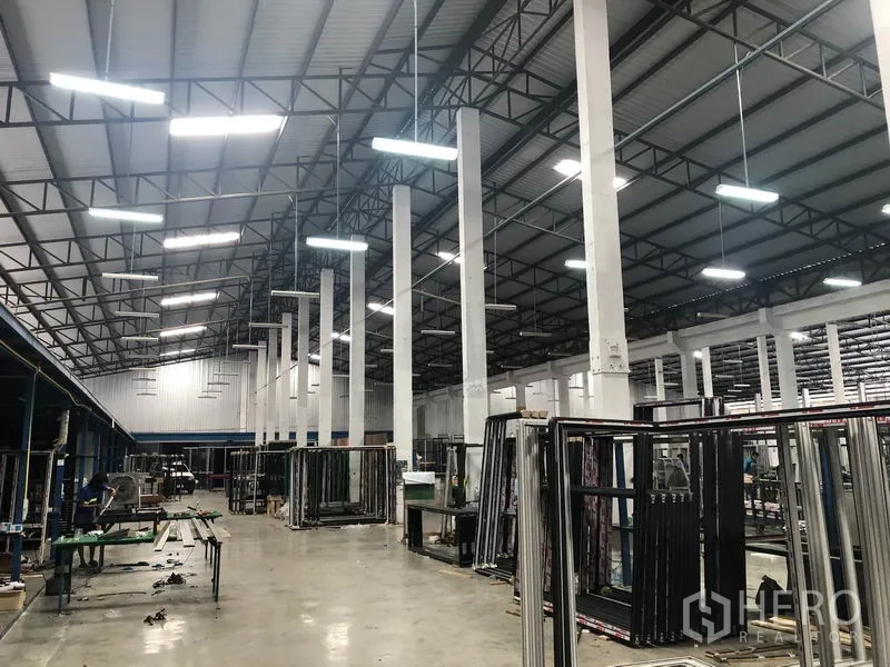 Warehouse for rent in Nonthaburi - Bright industrial hall showing steel trusses, suspended lights and organized work zones.