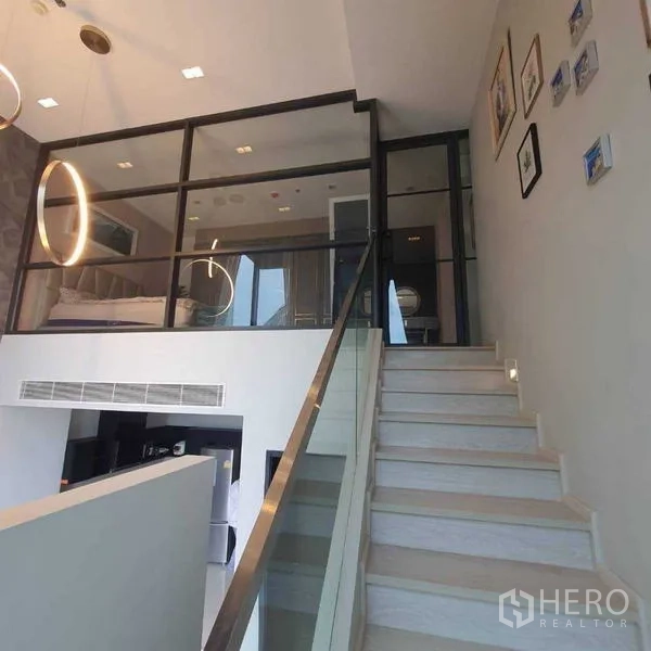 Condominium for rent in Khlong Toei, Bangkok - Staircase leading to a glass-walled mezzanine bedroom with circular pendant lights above.