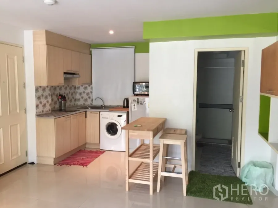 Condominium for rent in Khlong Toei, Bangkok - Compact L-shaped kitchen with light wood cabinets, floral backsplash, washing machine, and a small island with stools.