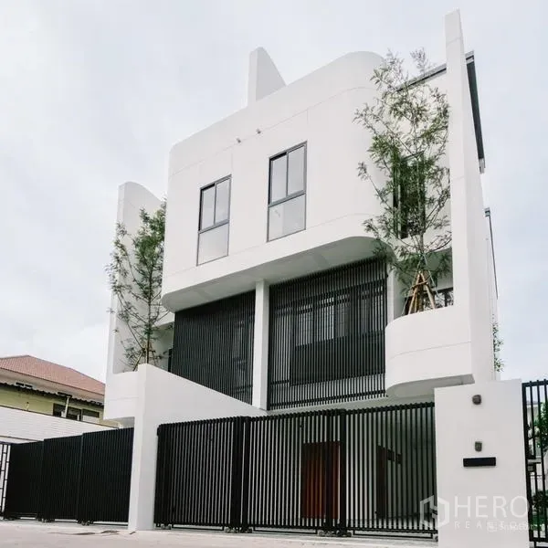 Townhouse for sale in Phra Khanong, Bangkok - Daytime view of the minimalist white townhouse facade with tall windows behind a black fence.