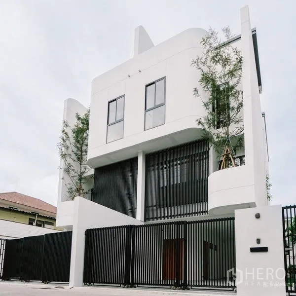 Townhouse for sale in Phra Khanong, Bangkok - Daytime view of the minimalist white townhouse facade with tall windows behind a black fence.