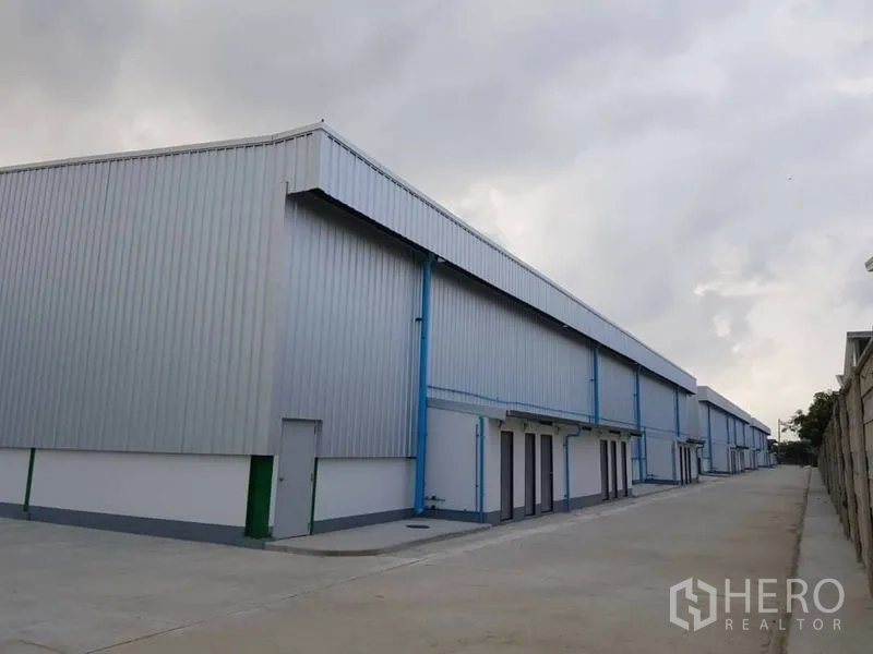 Warehouse for rent in Bang Phli, Samut Prakan - Row of metal-clad warehouse units with roller shutter doors and a paved driveway under cloudy skies.