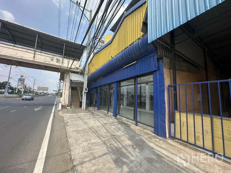 货栈 出租在Pak Kret, Nonthaburi - 临街仓库外观，黄色与蓝色立面，整面玻璃展示窗，旁有人行天桥。
