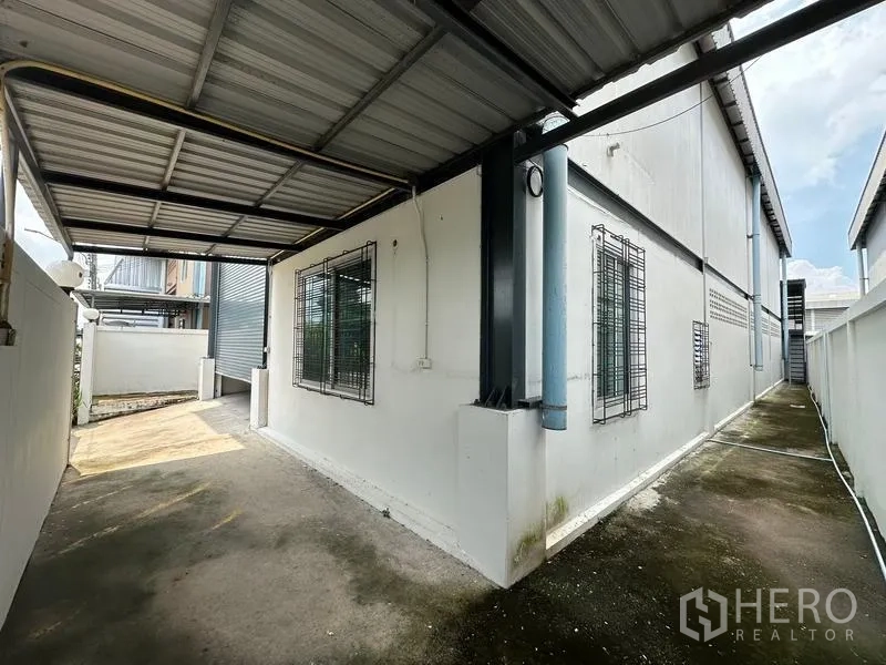 Factory for rent in Bang Bua Thong, Nonthaburi - Covered side yard with metal roof and barred windows along the factory wall for secure service access.