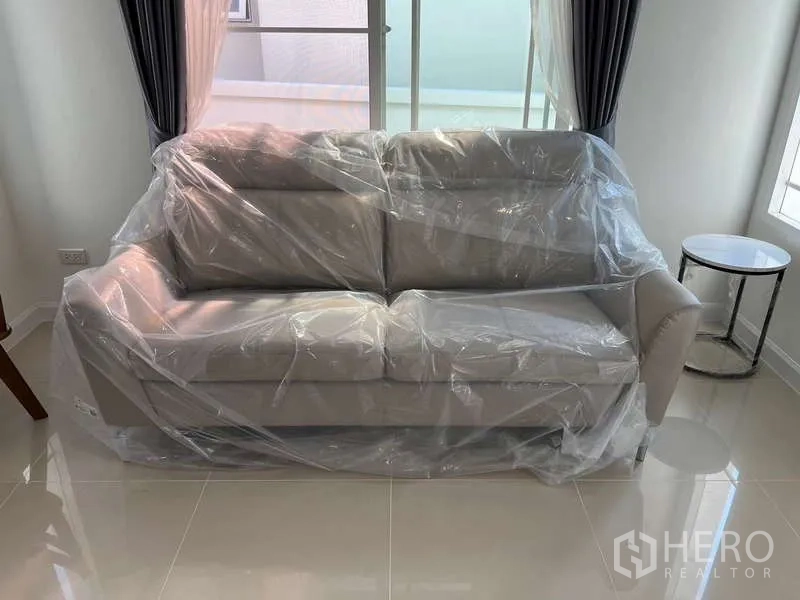 Detached House for rent in Bang Kapi, Bangkok - New grey sofa wrapped in plastic in a bright tiled living room.