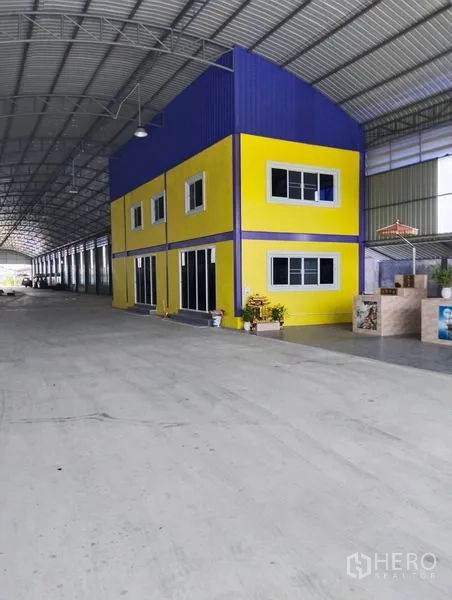 Warehouse for sale in Sai Noi, Nonthaburi - Spacious warehouse interior with high-arched steel roof and a two-storey yellow office block.