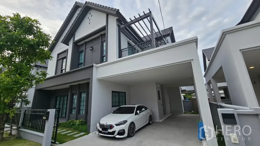 Detached House for rent or sale in Bang Phli, Samut Prakan - Modern two-storey detached house exterior with covered carport, white sedan, balcony pergola and landscaped front yard.