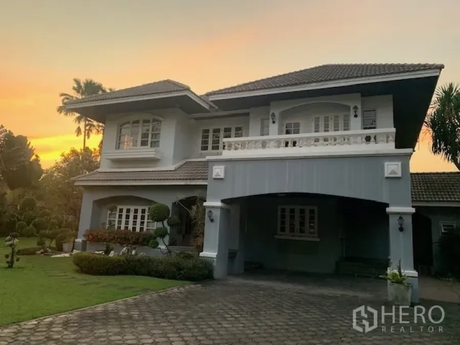 Detached House for rent in Khan Na Yao, Bangkok - Two-storey detached house exterior with covered carport, balcony, and large front yard at dusk.