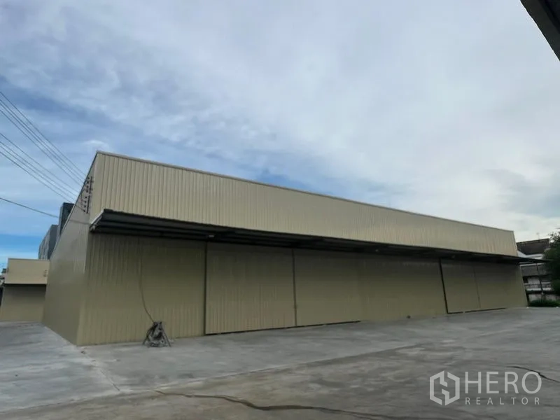 Warehouse for rent in Phra Khanong, Bangkok - Steel-clad beige warehouse with long sliding doors and a wide concrete yard under a cloudy sky in Phra Khanong, Bangkok.