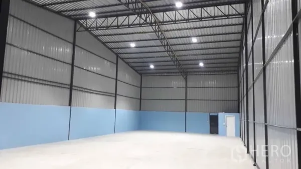 Warehouse for rent in Pak Kret, Nonthaburi - Open-plan warehouse bay with high ceiling, blue lower walls, large sliding doors, and a personnel access door.