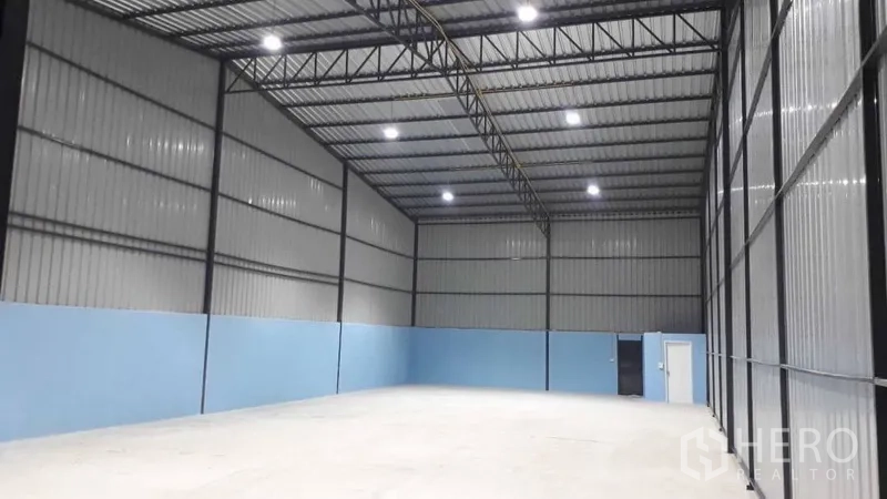 Warehouse for rent in Pak Kret, Nonthaburi - Open-plan warehouse bay with high ceiling, blue lower walls, large sliding doors, and a personnel access door.