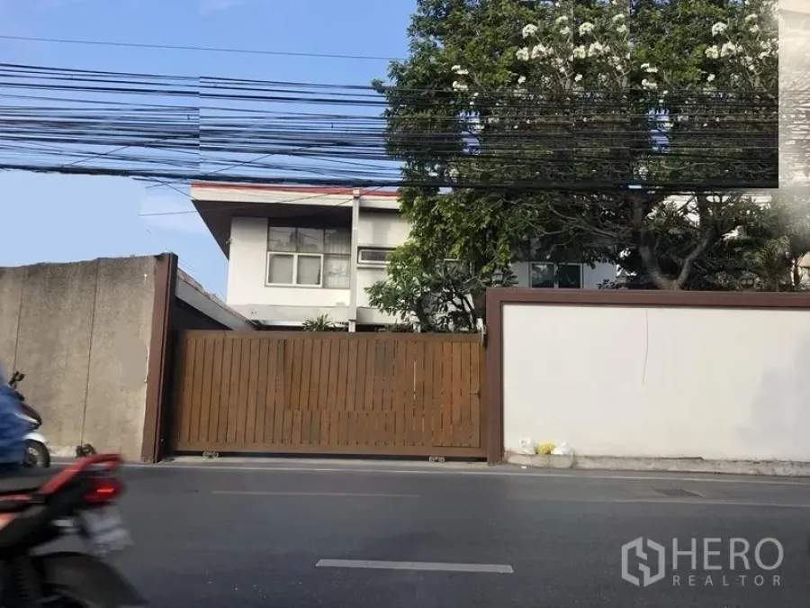 Retail Space for rent in Watthana, Bangkok - Street-facing gated retail compound with white boundary wall, wooden sliding gate, mature tree and two-storey building behind in Ekkamai, Bangkok.