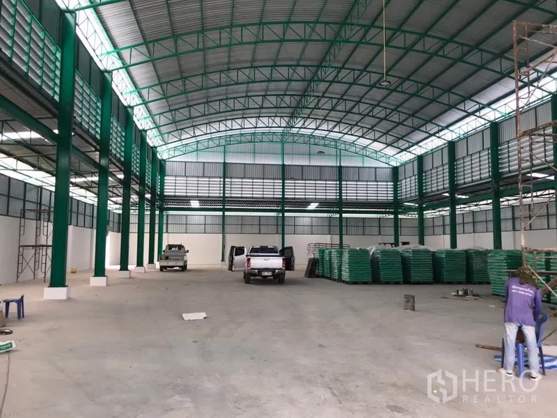 工厂 出租或出售在Mueang Samut Sakhon, Samut Sakhon - 开放式生产区，拱形屋顶下停有车辆与堆放物料。