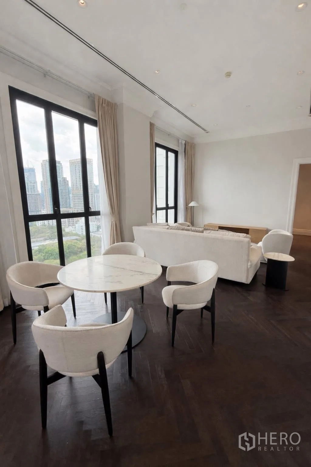 Condominium for rent or sale in Pathum Wan, Bangkok - Open living and dining area with a round marble table, sofa set, and skyline views through tall windows.