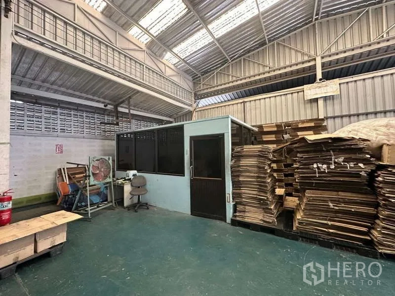 Warehouse for rent in Yan Nawa, Bangkok - Small onsite office cabin inside the warehouse beside stacked cardboard and equipment under a high roof.