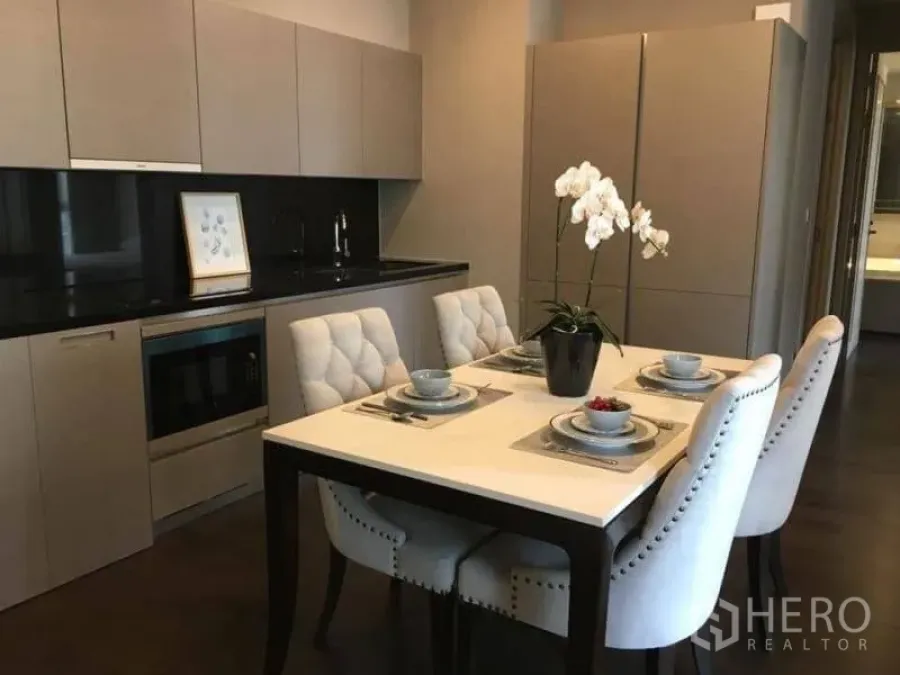 Condominium for rent in Watthana, Bangkok - Modern kitchen with black stone backsplash, built-in microwave, and a four-seat dining table with upholstered chairs and orchid centerpiece.