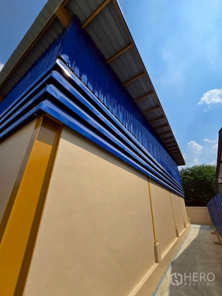 Warehouse for rent in Khlong Sam Wa, Bangkok - Side exterior of a warehouse with blue corrugated cladding, beige wall, yellow columns, and a clean driveway.